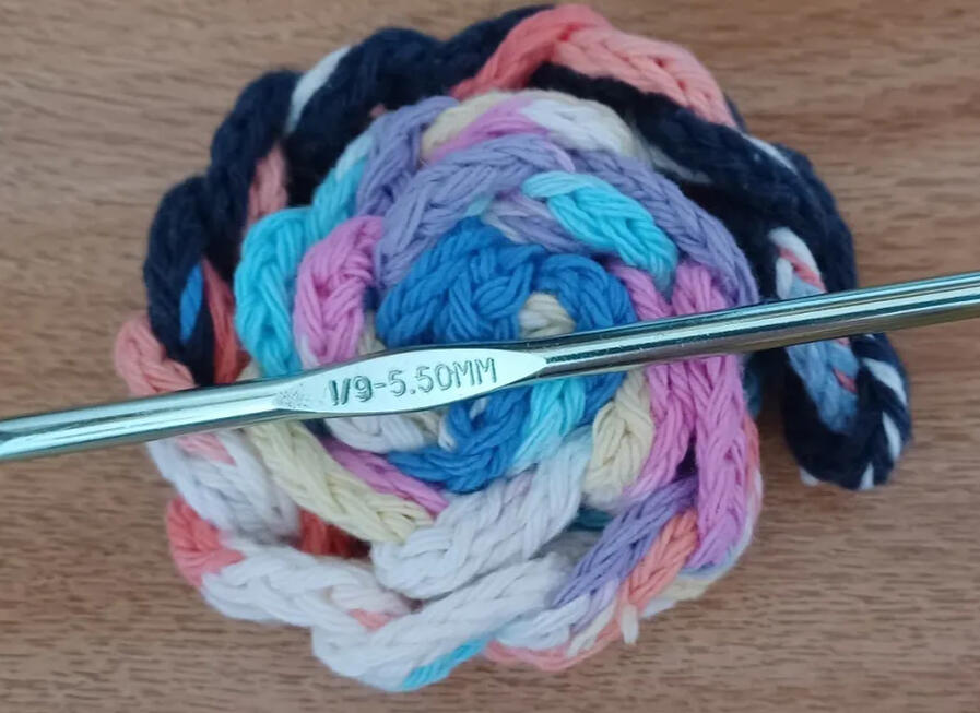 Braided i-Cord + hook a braided, crochet i-cord is wrapped into a colorful spiral with the crochet hook used to make the i-cord lying across the top of it. The background is my wooden desk.