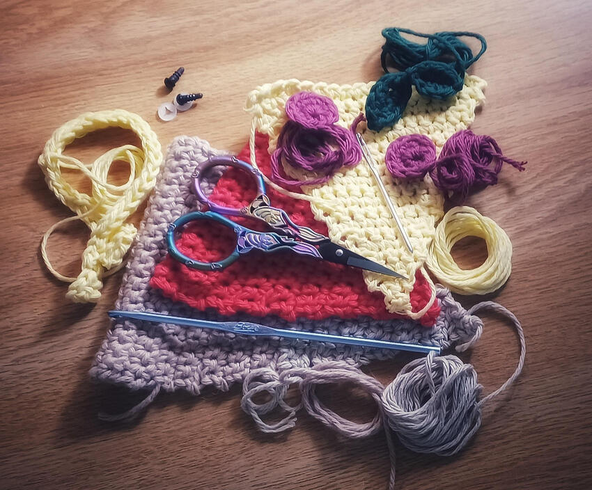 The pieces of a pizza amigurumi [crochet toy] await assembly. They are lying in an aesthetically staged heap of crochet shapes with the yarn ends still attached and wrapped in tight circles. Underneath the pile is the honey-golden wood of my desk.