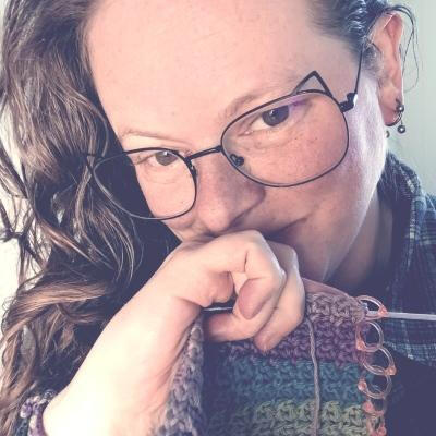 Laura | Lala Weiller, the site owner Laura Weiller, the owner and author of this site faces the camera. She is wearing a pair of glasses, the rims of which have cat ears on them. There's some partially crocheted, multi-colored yarn with stitch markers down the edge of it in her left hand, whi