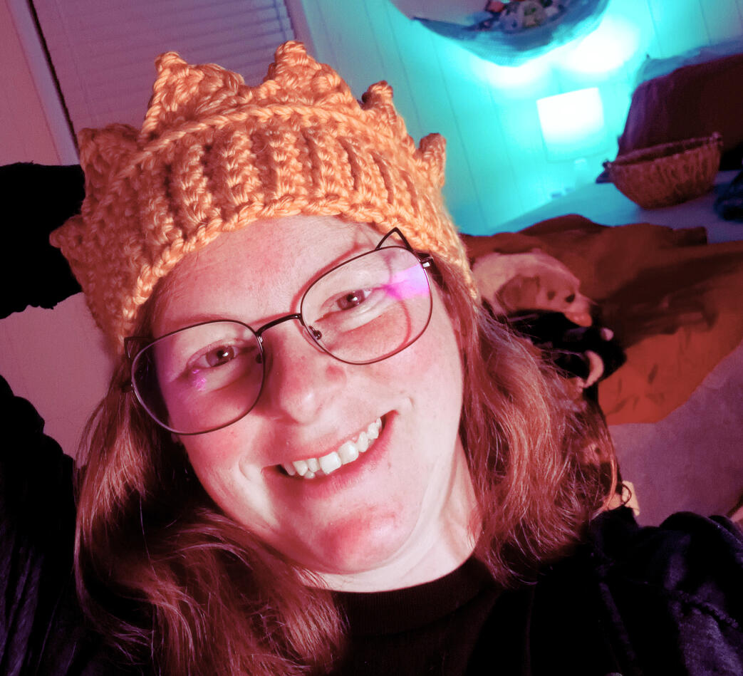 Laura Weiller, the owner and author of this site faces the camera. She is wearing a pair of glasses, the rims of which have cat ears on them. There's some partially crocheted, multi-colored yarn with stitch markers down the edge of it in her left hand, whi